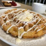Golden-brown crispy funnel cake dusted with powdered sugar, served on a white plate with a drizzle of chocolate sauce and fresh strawberries on the side.