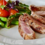Sliced medium-rare Denver steak served with fresh asparagus and cherry tomatoes on a white plate